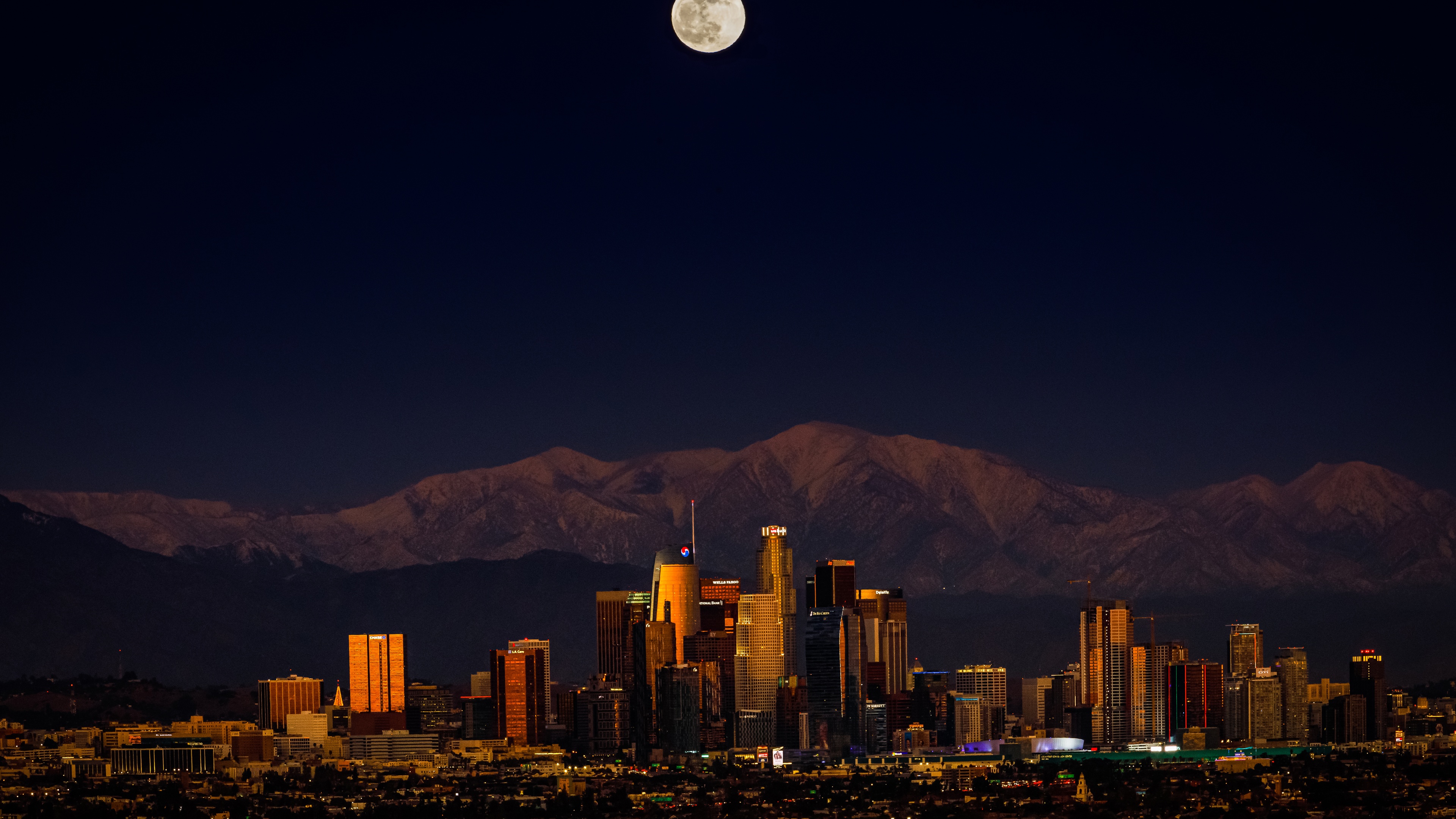A panoramic night view of the Los Angeles downtown cityscape featuring a large full moon in a dark sky.