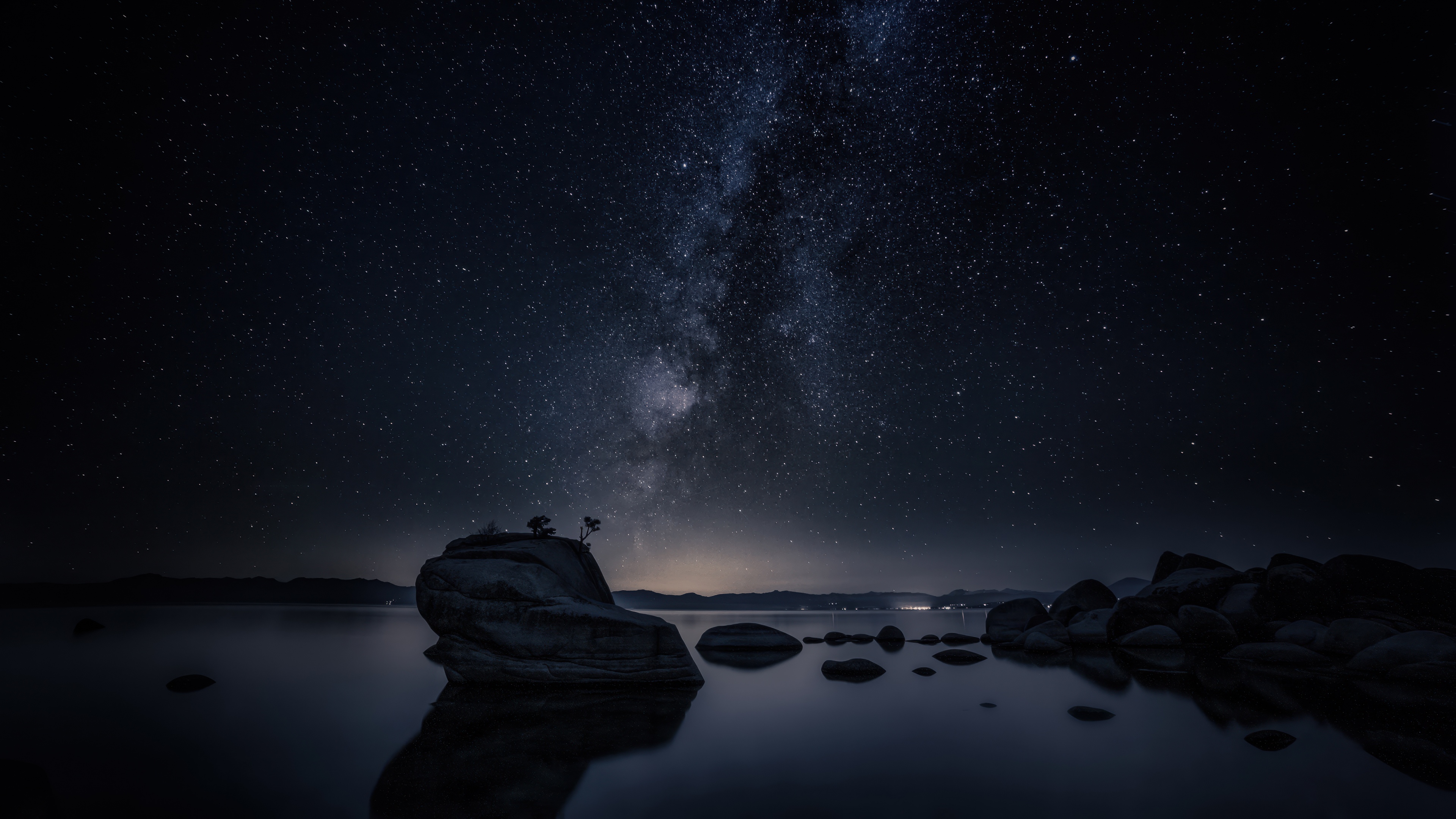 A wide-angle long exposure shot of the Milky Way galaxy and starry night sky reflecting in the still waters of Lake Tahoe with dark rock formations in the foreground.