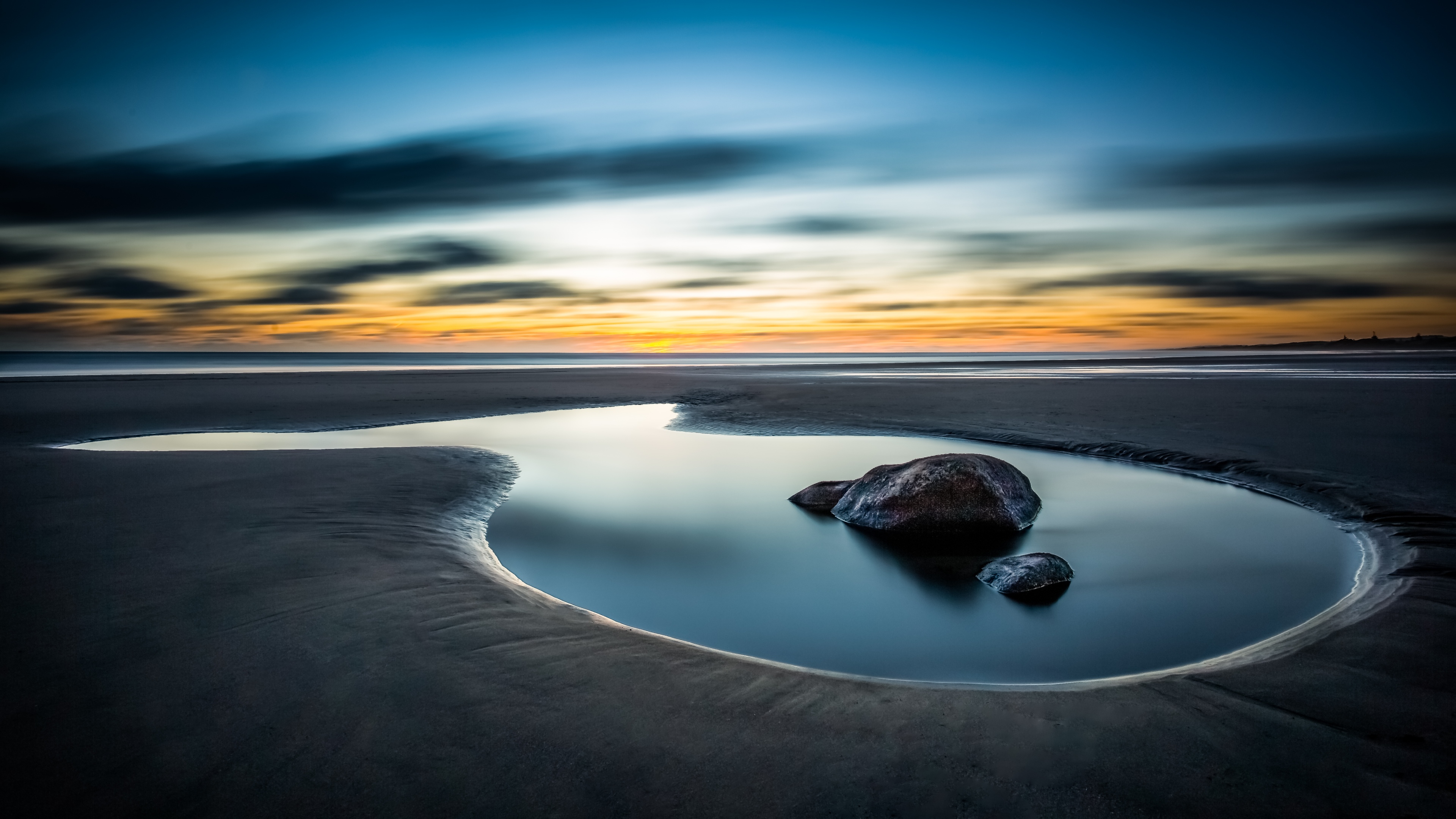 A tranquil beach landscape at sunset with smooth, misty ocean waves and golden reflections on the coastal shore under a vast sky.