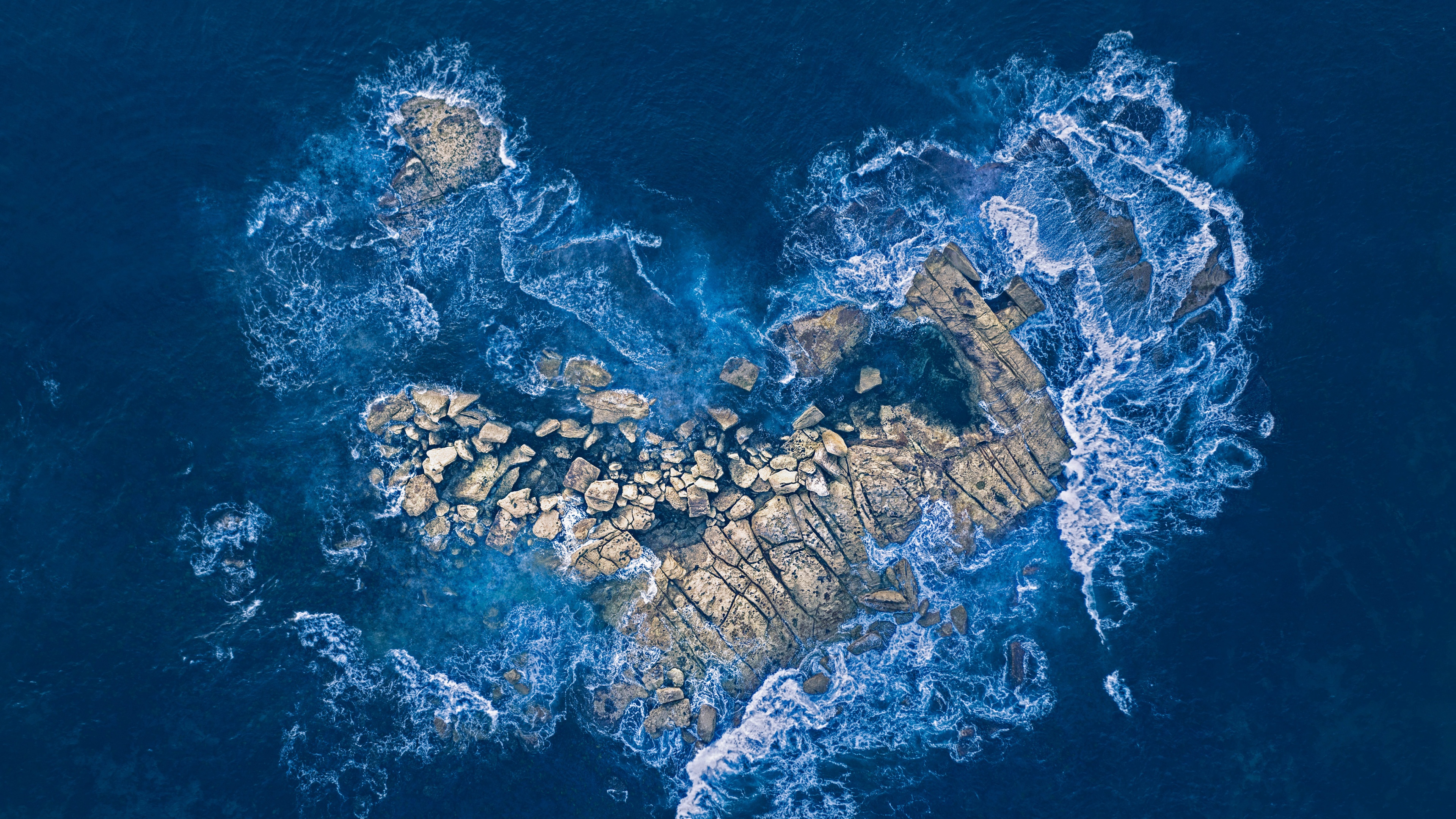 Aerial view of a heart-shaped rock island nestled in the middle of deep blue ocean waves.